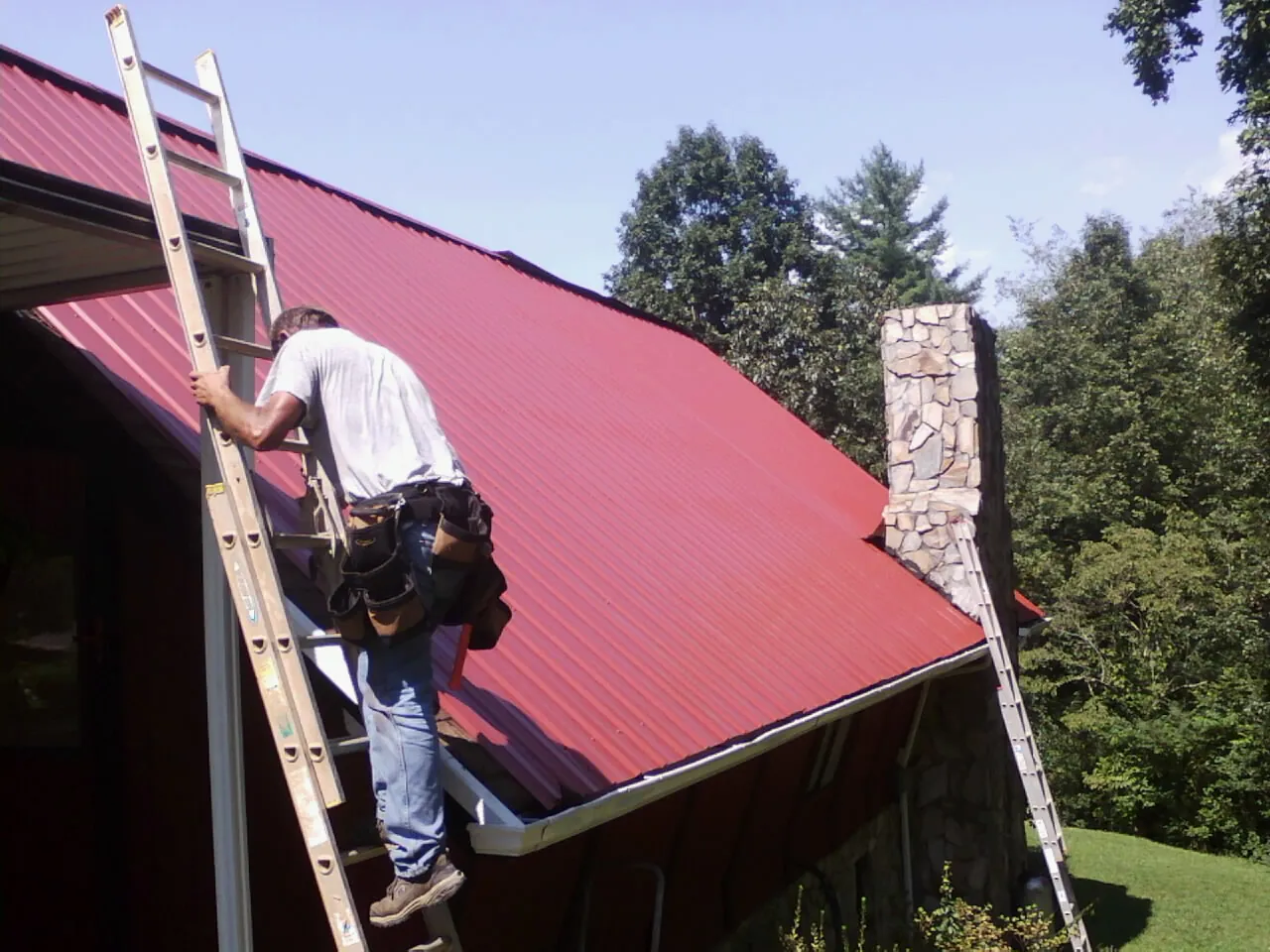 Roofer on a ladder beside a red metal roof and stone chimney — Soffit & Fascia Repair by Roofing Brothers in Brigham City