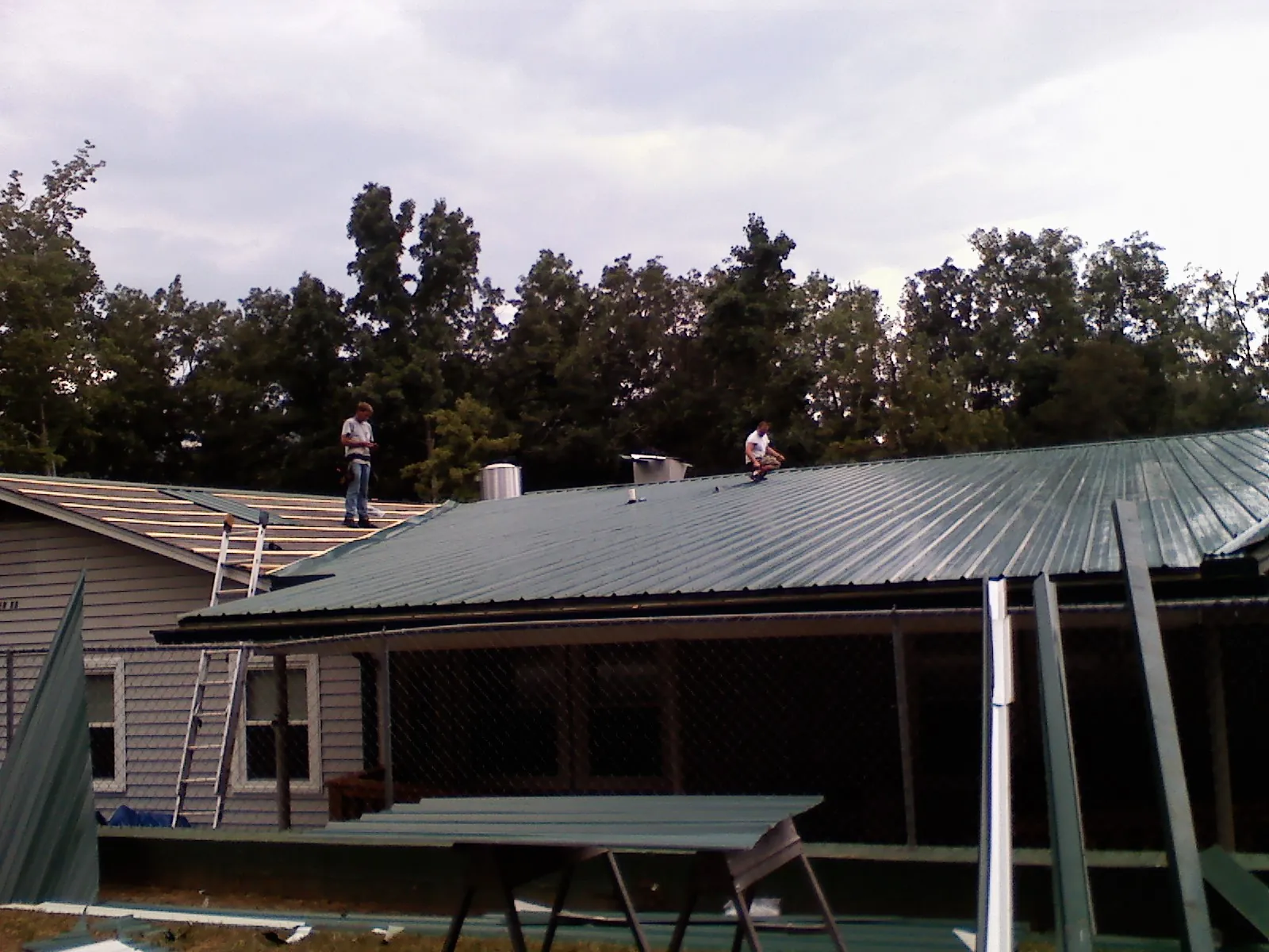 Two roofers installing green metal panels above a covered porch in Brigham City